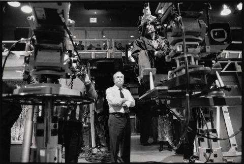 Lorne Michaels Collection Black and white. A man in a dress shirt and tie stands with his arms folded behind two giant cameras in a television studio.