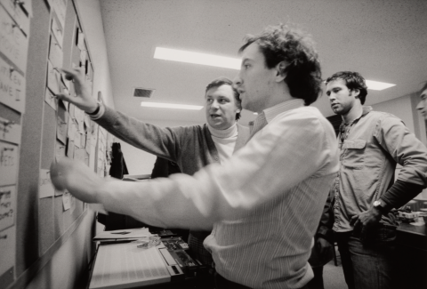 Lorne Michaels Chevy Chase Dave Wilson Black and white. In an office, three men rearrange a cork board full of notecards.