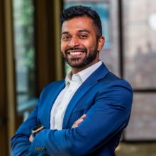 Gowtham Velu Gowtham Velu smiles with his arms folded in front of a blurred outdoor background. He wears a bright blue suit with a white button-up shirt, and a SmartWatch. In the background are two floor-to-ceiling windows, through which we can see an obscured building, a bright blue square, and a green tree.