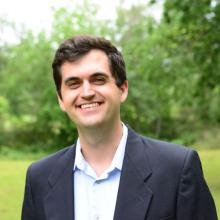 Cole Maguire Cole Maguire smiles at the camera wearing a black blazer and a light blue button-down shirt. He stands in front of a blurred background of green foliage and bright green grass.