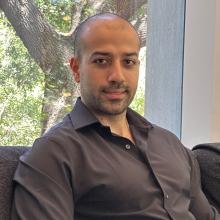 Khashayar Moshksayan sits on a black couch, wearing a brown button-up shirt. Behind him is a white wall with a window. Through the window are bright green leaves and the thick trunk and branches of a tree.