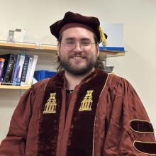 Keegan Lorenz-Ochoa wears the doctoral regalia for the University of Texas at Austin. The regalia is dark red, with a brown velvet sash around the neck which has the tower embroidered along both sides, in gold. He also wears a dark red velvet cap with a gold tassel, and glasses. Lorenz-Ochoa stands in front of a bookshelf of chemistry books, in a white room.