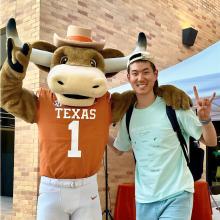 Yanbing Zhou wears a teal t-shirt, black backpack, light blue shorts, and a beige baseball cap turned backwards. Next to him is Hook'em, a mascot of a brown bull with white and black horns, an orange Texas jersey, white shorts, and a beige cowboy hat. Both Hook'em and Yanbing smile and make the "Hook 'em" hand sign in front of a beige brick building. Behind them is a light blue tent over a table draped in red cloth.