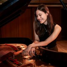 Ellen Sirower smiles and looks down into the inside of a grand piano. She wears a short-sleeved black shirt, brown pants, and a silver necklace. Her hands are crossed over the copper inside of the piano soundboard.