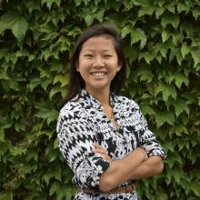 Emma Palmer smiles with her arms crossed in front of a hedge of lush green leaves. She wears a black and white patterned button-down shirt, white pearl earrings, a gold necklace, and a braided brown belt.