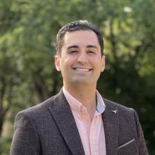 Iman Salafian Iman Salafian smiles in front of a green forest background, wearing a grey suit jacket over a pink button-down shirt.