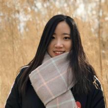 Zexi Zhou 2 Zexi Zhou wears a black jacket and a thick grey scarf with baby blue and pink stripes. She smiles in front of a forest with light orange leaves.