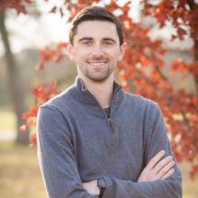 Nathan Herrmann Nathan Herrmann wears a grey sweater and smiles with his arms crossed in front of a tree with orange autumn leaves.