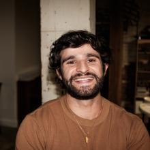 Darius Atefat-Peckham Darius wears a brown sweater and a gold necklace and smiles in front of a white pillar.