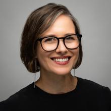 A woman wear a black blouse and black glasses with long silver earrings. She stands in front of a grey background.