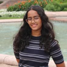Ishita Chitturi A woman with long dark hair wears glasses and a black and grey striped sweater in front of a large stone water fountain.