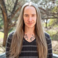 Draconis von Trapp A man wears a brown-and-grey striped sweater in front of a blurred forest background.