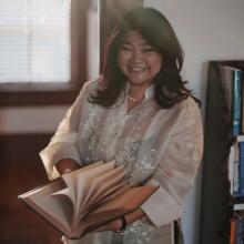 Anna Marinela Lopez A woman wears a white blouse and flips the pages of a book. She poses in front of a bookcase and a window.