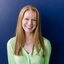 Suzie Wright A woman wears a lime green cardigan and a gold necklace and smiles in front of a dark blue background.