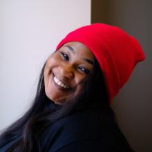 Annase Raji A woman wears a dark sweater and a red beanie in front of a white wall. Her head is tilted and she smiles at the camera.