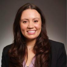 A woman smiles in front of a grey background, wearing a black blazer and a purple blouse.