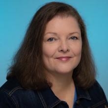 A woman smiles in front of a teal background, wearing a dark blue jean jacket.