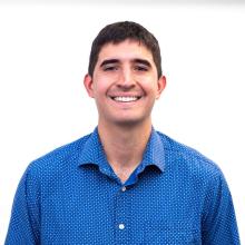 A man in a blue shirt smiles in front of a white background.
