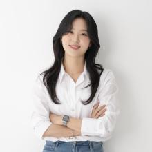 A woman wears a white blouse and jeans in front of an off-white background.