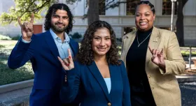 Ashley Alvarado, Ali Altahafi, and Kennedy Simon make the "Hook Em Horns" hand sign in front of a building on the UT-Austin campus.