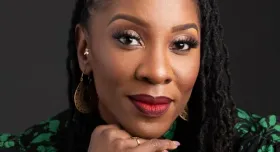 Aysha Upchurch wears a green and black floral shirt in front of a black background. Her head rests on her hand.