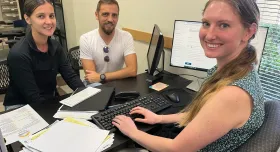 A woman sits at a desk full of tax forms and a keyboard. In front of her are a man and a woman. All three of them smile at the camera.