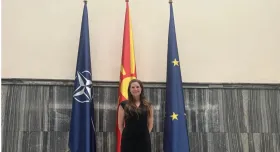 A woman in a black dress stands in front of three Macedonian flags inside a building in North Macedonia.