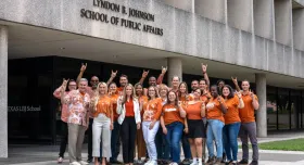 A group of students and faculty wear orange University of Texas at Austin shirts and make the Hook 'em horns sign in front of the Lyndon B. Johnson School of Public Affairs building.