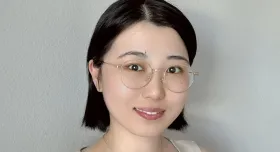 A woman wears glasses, a gold necklace, and a beige shirt and stands in front of a white wall.