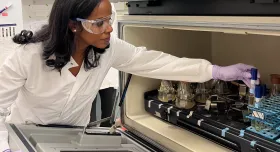 A woman wears clear safety glasses, a white lab coat, and purple gloves as she opens up a storage space, resembling an oven, for test tubes.