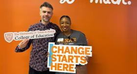 Two people stand in front of an orange wall painted with letters reading "I love coe so much." They both hold signage for the UT College of Education