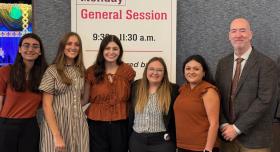 Five students and one professor wear business casual and stand ini front of a banner advertising the General Session.