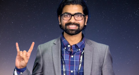 A man wears a suit and makes a Hook 'em sign in front of a galaxy background.