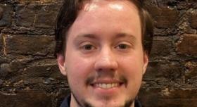 A person smiles in front of a brown brick wall.