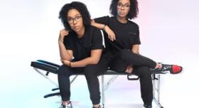 Twins in black clothes sit next to each other on a massage table against a white background.