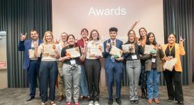 Eleven students in businesswear pose with their award certificates at the front of a conference room.