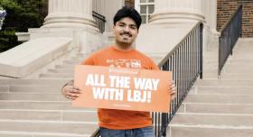 Caleb Garcia stands on some outdoor steps with an orange sign reading "All the Way with LBJ!"