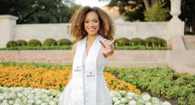 Chanel Williams wears a white convocation sash and makes a Hook 'em Horns hand sign in front of a flowerbed on the UT Austin campus.