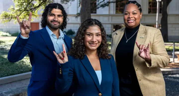 Ashley Alvarado, Ali Altahafi, and Kennedy Simon make the "Hook Em Horns" hand sign in front of a building on the UT-Austin campus.