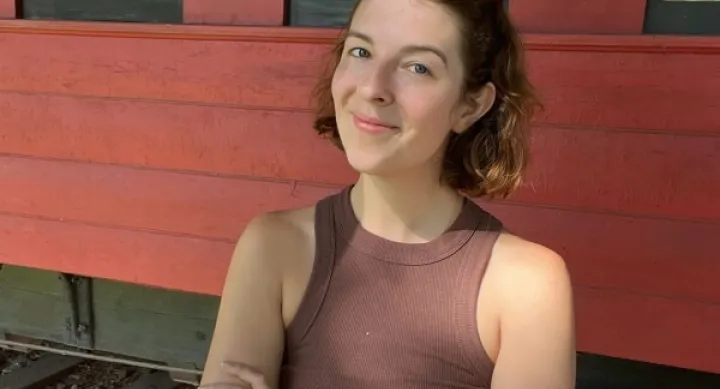 Gwen Ruth Jones wears a brown top and smiles in front of a red building with her arms folded.
