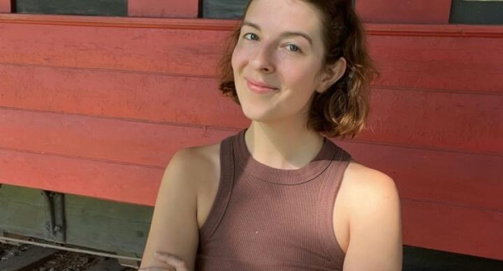 Gwen Ruth Jones wears a brown top and smiles in front of a red building with her arms folded.