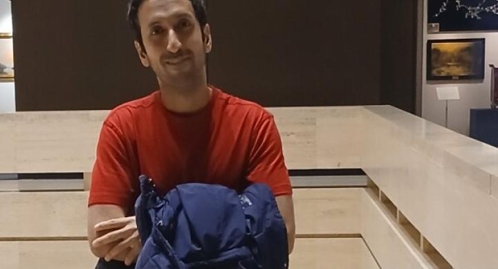 Pouya Neokoeui wears a red shirt and holds a blue winter coat inside a museum with black walls and paintings in the background.