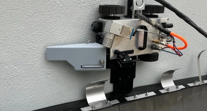 A metal contraption rests against a white plaster wall. The contraption has two black knobs at the top, and black and orange wires coming out of the side.