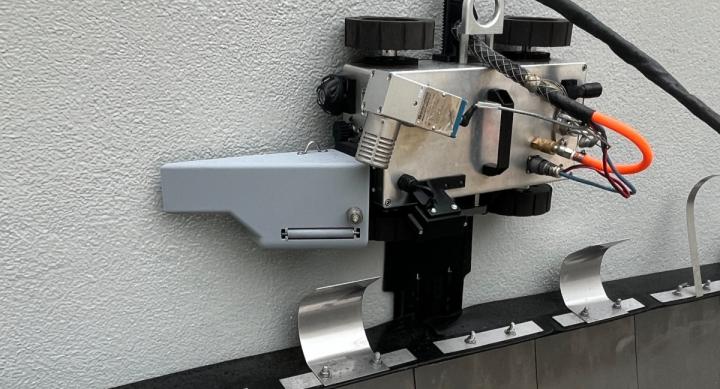 A metal contraption rests against a white plaster wall. The contraption has two black knobs at the top, and black and orange wires coming out of the side.