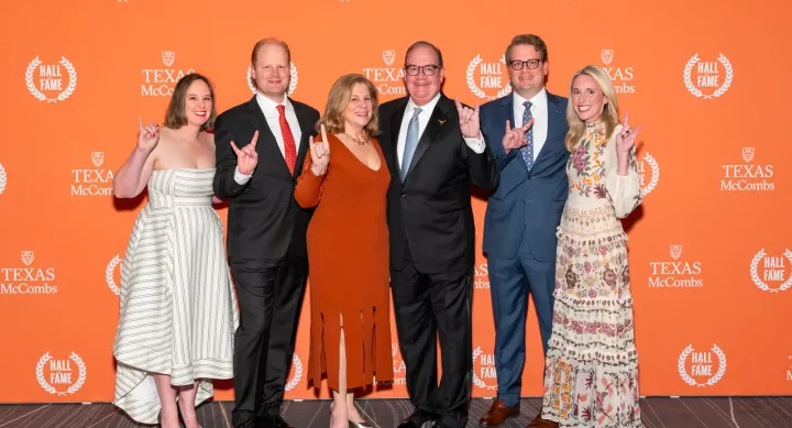 Six people stand in front of an orange background covered in logos for the Texas McCombs Hall of Fame. All of the people make the "hook 'em horns" sign with their hands. The people are dressed as follows, from left to right: a woman in a white and green striped dress, a man in a black suit with a red tie, a woman in a red dress, a man in a black suit and blue tie, a man in a blue suit and blue patterned tie, and a woman in a cream colored, floral patterned dress.