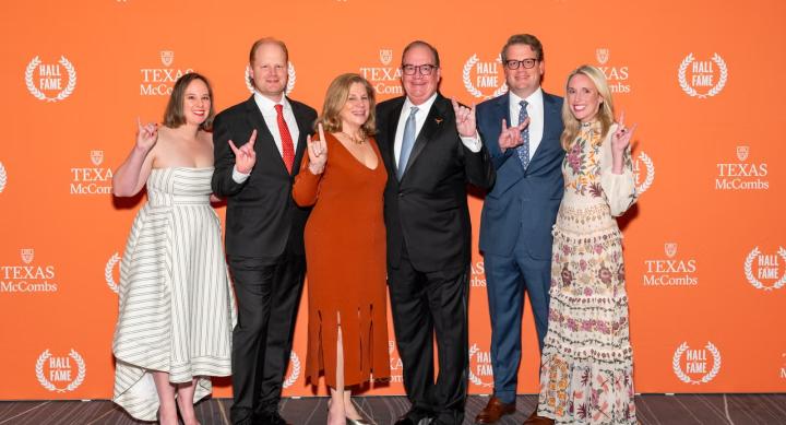 Six people stand in front of an orange background covered in logos for the Texas McCombs Hall of Fame. All of the people make the "hook 'em horns" sign with their hands. The people are dressed as follows, from left to right: a woman in a white and green striped dress, a man in a black suit with a red tie, a woman in a red dress, a man in a black suit and blue tie, a man in a blue suit and blue patterned tie, and a woman in a cream colored, floral patterned dress.