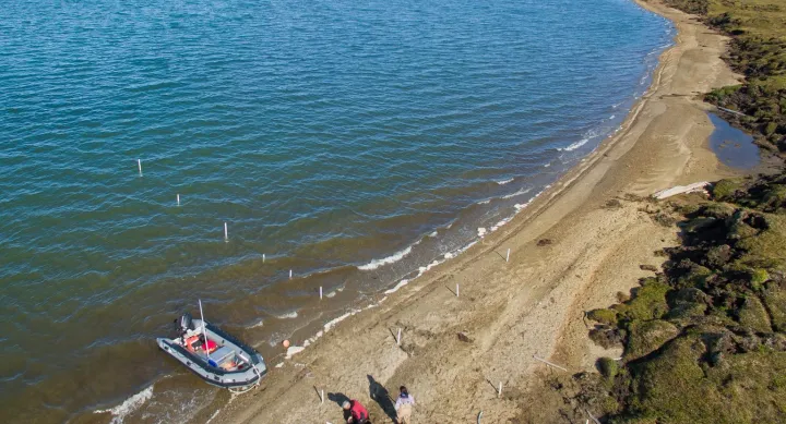 An aerial image of the study site in Kaktovik Lagoon in Northern Alaska. A small blue boat pulls up along the beach while two people in red life jackets and a person in beige overalls stand next to black boxes of equipment on the shore.
