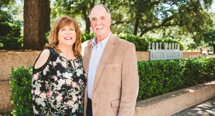 George and Barbra Polansky smile in front of a forest background. Behind them is a brown stone wall lined with a hedge, and a grey sign reading "Fort Worth Water Gardens." George wears a brown suit jacket and light blue shirt. Barbra wears a black blouse with yellow and pink flowers and green leaves.