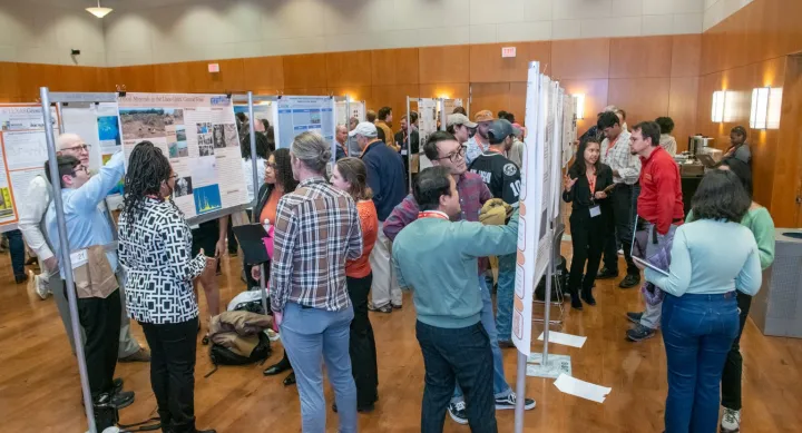 University students and professors stand in a room with wood-paneled walls and hardwood floors, all crowding posters hung on metal stands and lined in rows throughout the room. The students presenting their posters wear red lanyards.