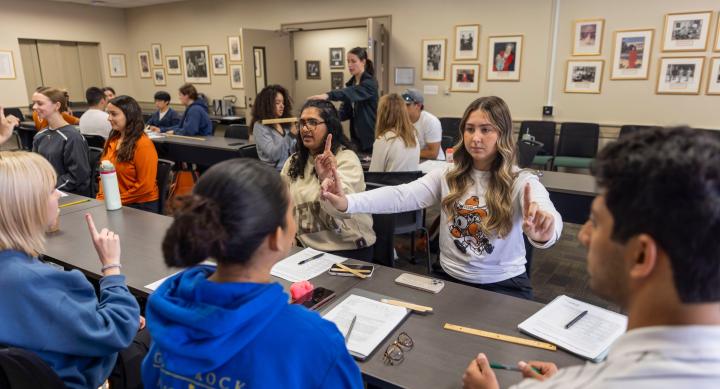 Moody students sit around tables in the Lady Bird Johnson conference room. They hold up fingers in the air as they test their vestibular sense. There are notebooks, pens, and rulers on the table in front of them.